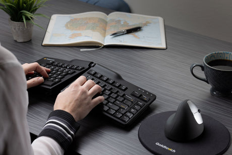A woman typing on a keyboard at her workstation, also featuring a Semi-Vertical Mouse and mouse pad. Her desk also includes a plant, planner with pen, and cup of coffee.
