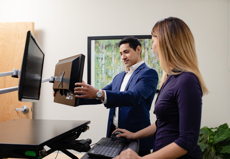 Woman sharing her screen with a male coworker who is easily able to swivel the monitor into view on the EasyLift Monitor Arms, working at an EasyLift Desk, with a Go!2 Mobile Keyboard, and Semi-Vertical Mouse.