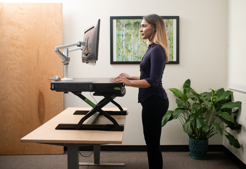 Female office worker looking at monitors mounted on a dual monitor arm, while using an EasyLift Desk from Goldtouch.