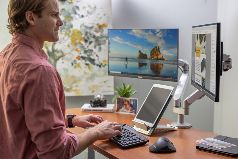 A man smiling while typing on a Goldtouch Elite Keyboard. He is looking at his monitor while typing. His desk has a book, camera, picture, Go! Travel Laptop Stand with iPad on it, Newtral 3 Mouse, iPad case.