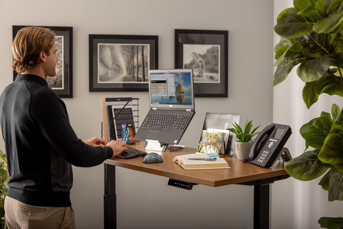 Male office worker at his workstation typing on a Goldtouch Go!2 Mobile Keyboard, with a FlexMouse, and Go! Travel Laptop and Tablet Stand raising his laptop up to eye level. The desk features various other things like a phone, book, pen, file holder, and fake plant. There is a plant in the corner and artwork on the wall.