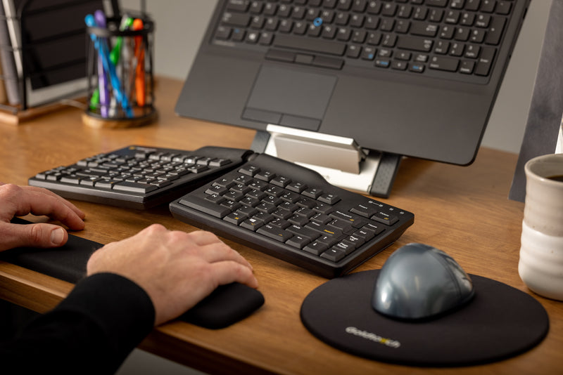 A closeup of a male office worker relaxing his hands on a wrist rest at his desk. The desk has his Goldtouch Elite Keyboard, FlexMouse, Round Mouse Pad, Go! Laptop and Tablet Stand with laptop on it.