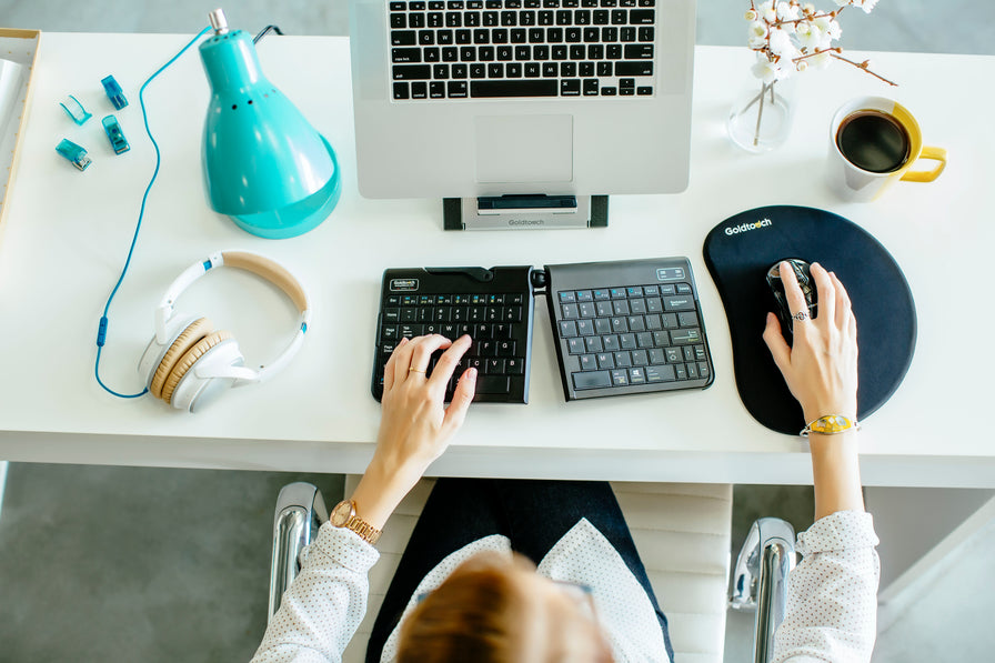 Female office worker sitting at a workstation, typing on a Goldtouch Go!2 Bluetooth Keyboard, mousing with an Ambidextrous Mouse, placed on a Mouse pad, looking at a laptop placed on a Go! Travel Laptop and Tablet stand with various office decor such as a coffee cup, headphones, and lamp.