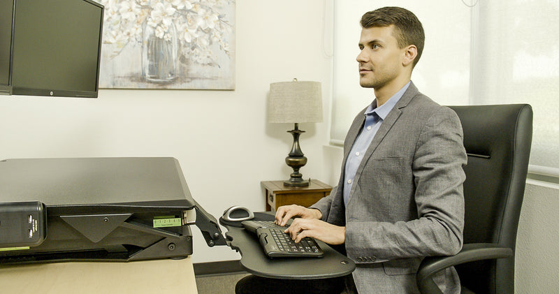 Man Working at Computer Workstation, including the Goldtouch EasyLift Desk with keyboard tray extended, Go!2 Mobile Keyboard, Bluetooth Comfort Mouse, and Round Mouse Pad.